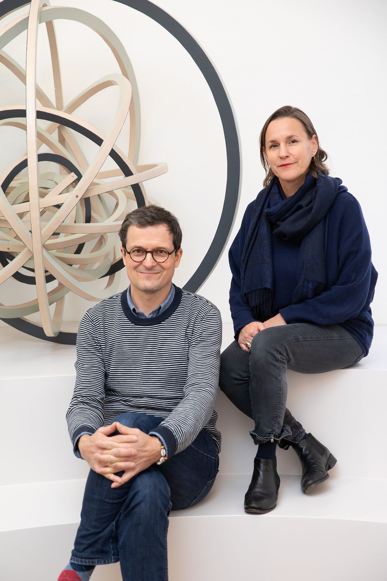 Zwei Perosnen sitzen in einem weißen raum, eine Frau mit braunem Haar und ein Mann mit Brille